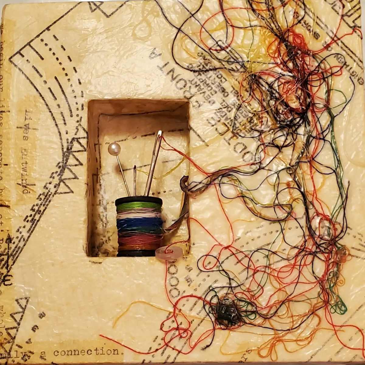 Lives Entwined by Jan Somers  Image: The shadow box is covered with dress patterns and creating a spool of multicolored threads. The poem is written on the side using a typewriter 