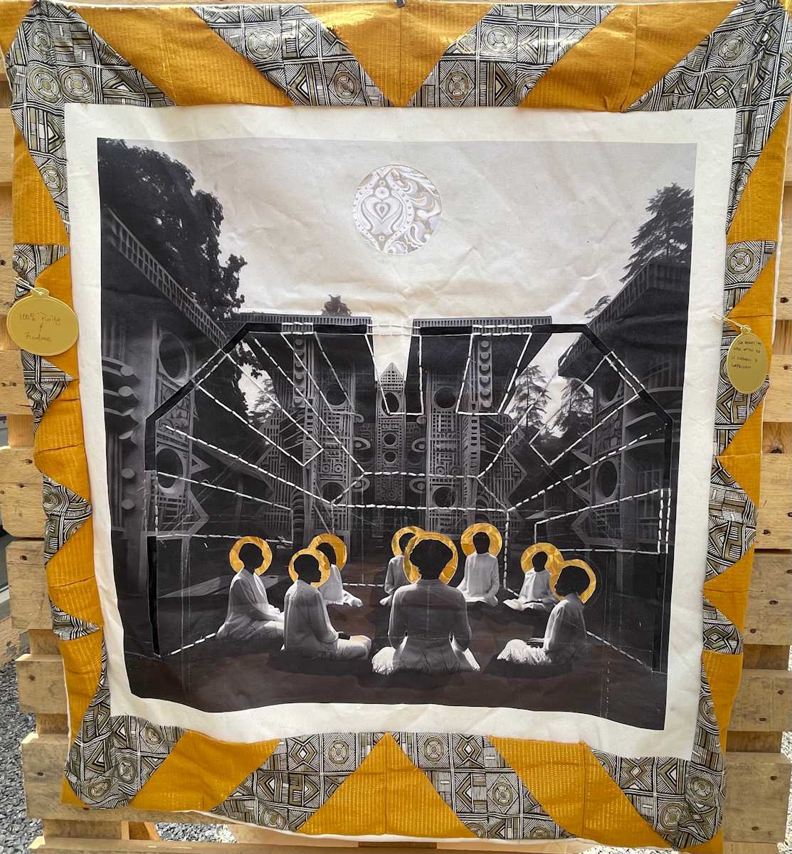 Nkpọchi Mmuo: Gateway to the Ancestors by Ifeoma Ebo  Image: In Igbo tradition, the nkpọchi mmuo (spirit gateway) is where the living commune with the ndichie to seek guidance and healing. This quilted ceremony depicts the sacred gathering known as igba ndu (consultation of life), where community members enter the ancestral realm through ritual and reverence. The eight figures completing your circle of nine, crowned with golden ichi markings, represent the sacred number in Igbo cosmology—symbolizing completion, wisdom, the spiritual emergence and the awakening of Chi. 
The architectural backdrop echoes the mbari houses of Igboland—sacred structures built to honor the earth goddess Ani and serve as bridges between worlds. These towering forms, rendered in deep shadows against the pale sky, create a threshold space where uwa (the visible world) meets ala mmuo (the spirit realm). The radiating lines connecting earth to sky represent the chi pathways that allow ancestral energy to flow into the community gathering.
The vibrant Ankara and Aso-Oke border cloth, with its interlocking geometric patterns, honors the Nigerian textile traditions while serving as akwa mgbochi (protective wrapping) around the sacred vision. These golden and gray fabrics mirror the colors of palm wine and kola nut—essential elements in Igbo ceremonies that invite ancestral presence. The textile choice transforms the quilt's edges into a ritual boundary, containing the spiritual energy within.
Above, the glistening moon suggests the watchful eye of Chukwu (the Supreme Being), while the earthbound figures demonstrate that through collective omenala (traditional practice), communities can access the infinite wisdom of those who have joined the ndichie. Here, ceremony becomes technology, and ancestral intelligence flows through the ritual space like light through darkness.