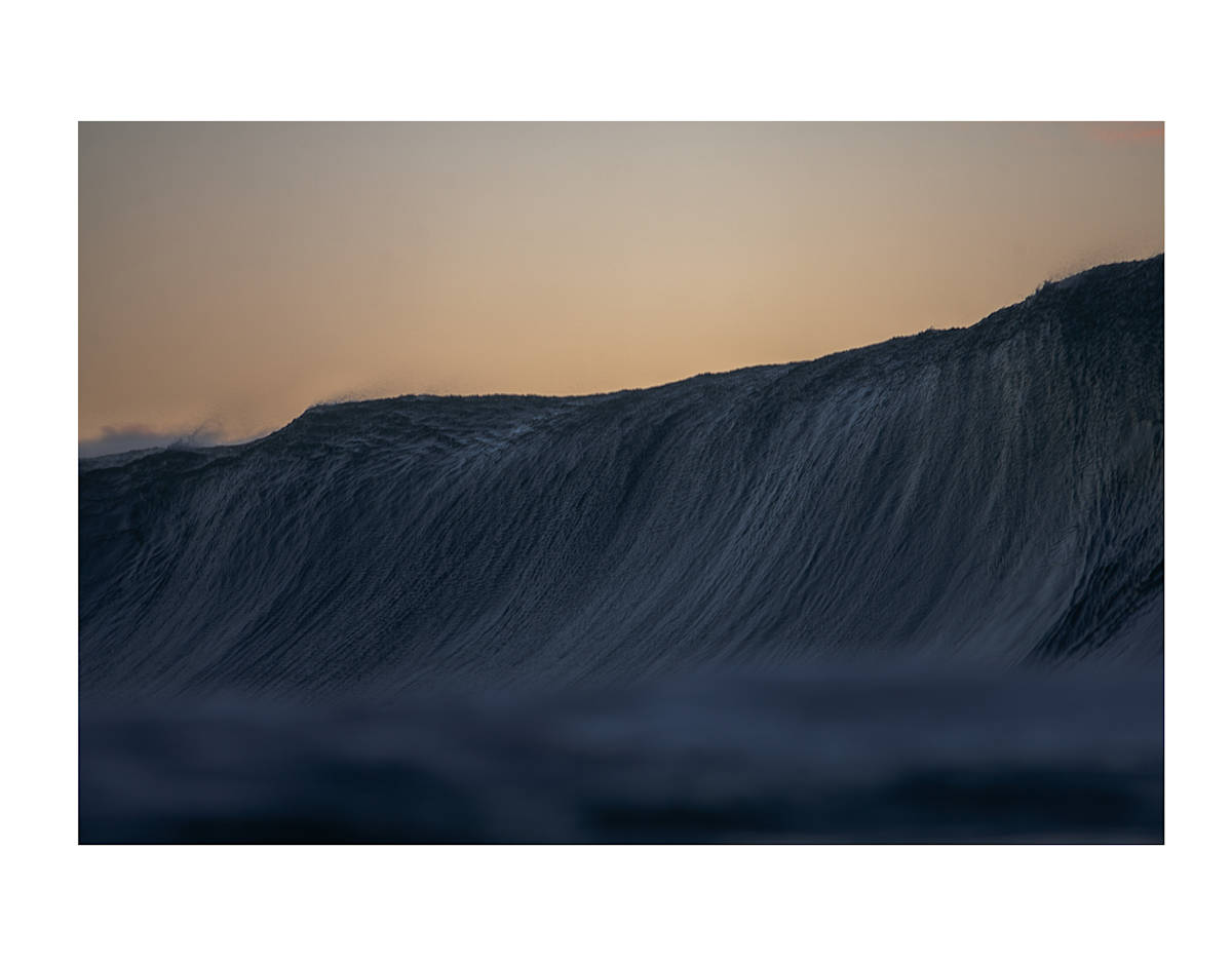 Verticle Pipeline by Daniel Russo  Image: On a late January afternoon, just after sunset, a rogue set of waves crashed onto First Reef Pipeline. I was the last person lingering in the water, eagerly hoping to capture the magic of an empty wave through my lens. The scene was breathtaking as the wall of water legged perfectly onto the first reef, forming an impressive vertical mountain of energy. The hues of the setting sun painted the sky with vibrant colors, adding to the drama of the moment. Sunset has always been my favorite time of day for documenting winter swells; the combination of the peaceful evening light and the powerful ocean creates a serene yet exhilarating experience.