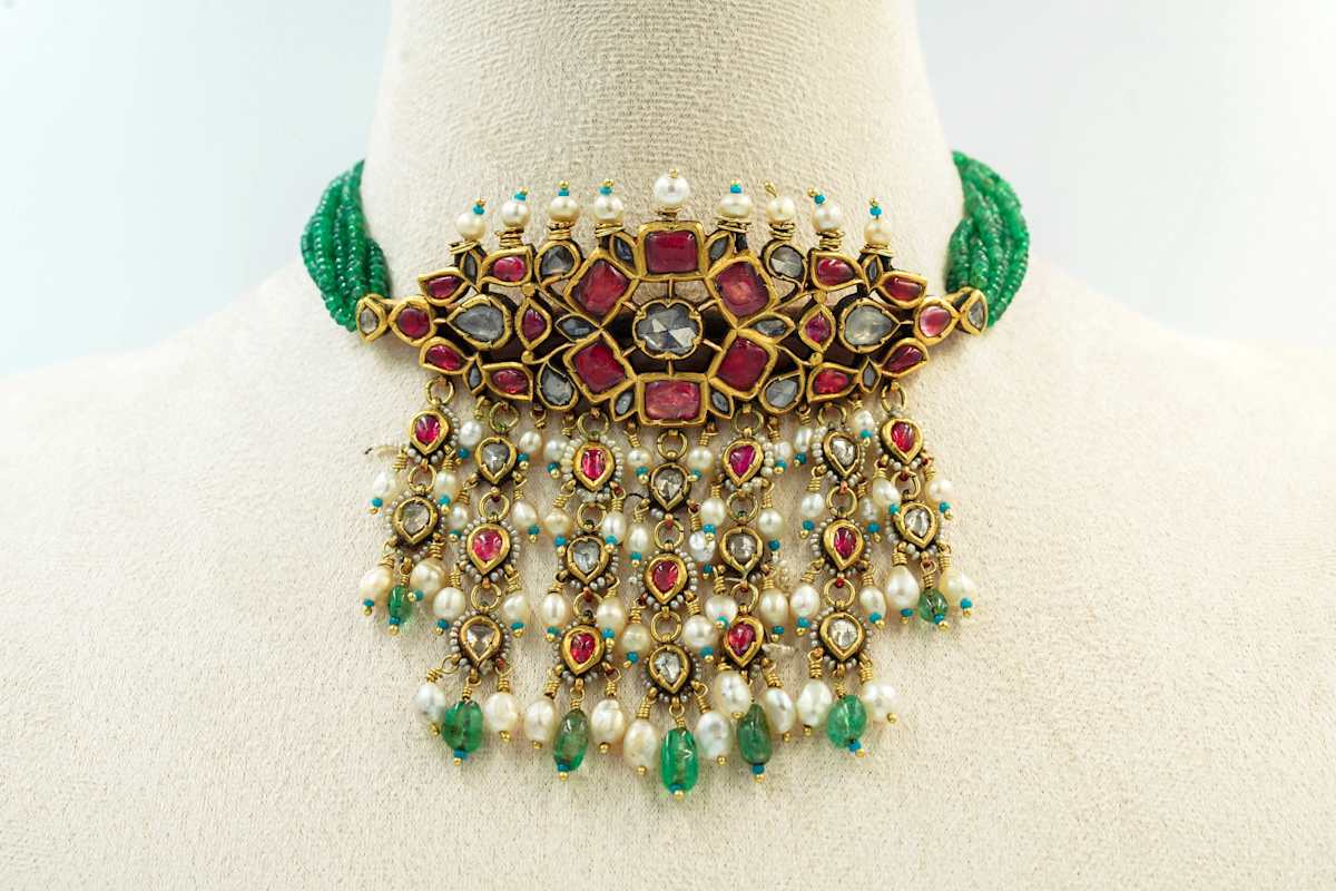Gold Choker  Image: Strung with multiple strands of emerald beads, this reversible choker features  diamonds, rubies, and pearls in kundan settings. Cascading fringes with ruby-centred teardrops and pearl-emerald drops enhance its movement. The reverse reveals intricate red and green enamel floral work, showcasing exceptional dual-sided craftsmanship.

