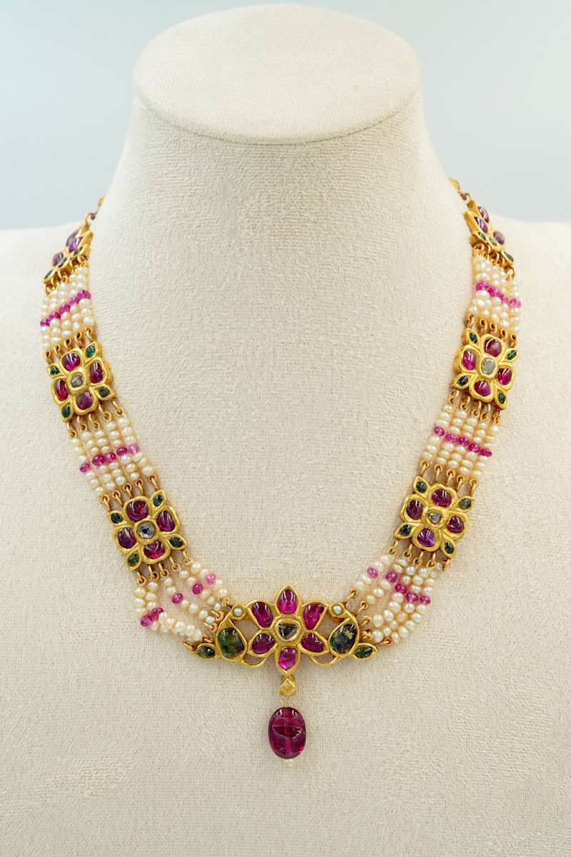 Necklace  Image: Composed of alternating strands of pearls and pink beads, this necklace is adorned with gold floral panels set in kundan technique, featuring rubies, emeralds, and uncut diamonds. A central floral motif with a dangling ruby bead adds a dramatic focal point.