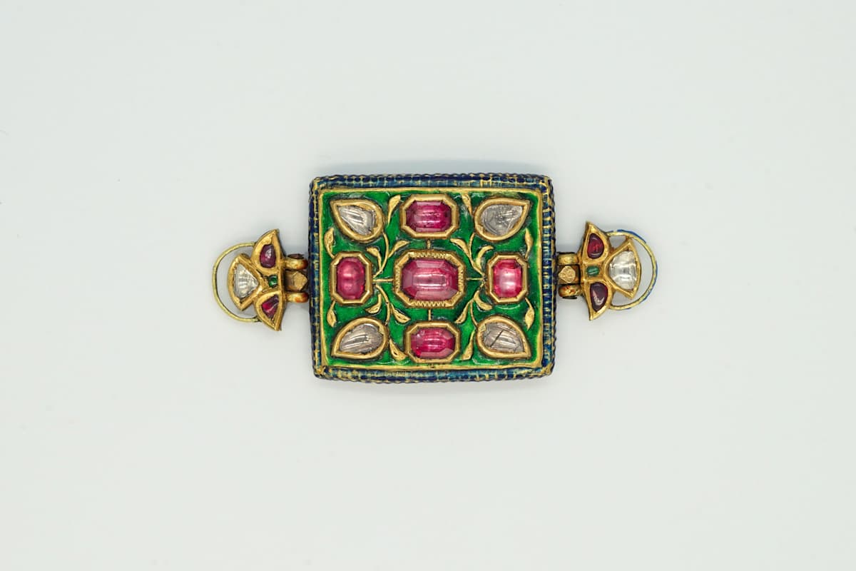 Gold armlet (Bazuband)  Image:  This regal bazuband showcases rubies and uncut diamonds in kundan settings against green enamel, echoing the precision of miniature painting. The reverse reveals vivid enamelled blooms on a deep blue ground, reflecting the dual artistry of the piece.
