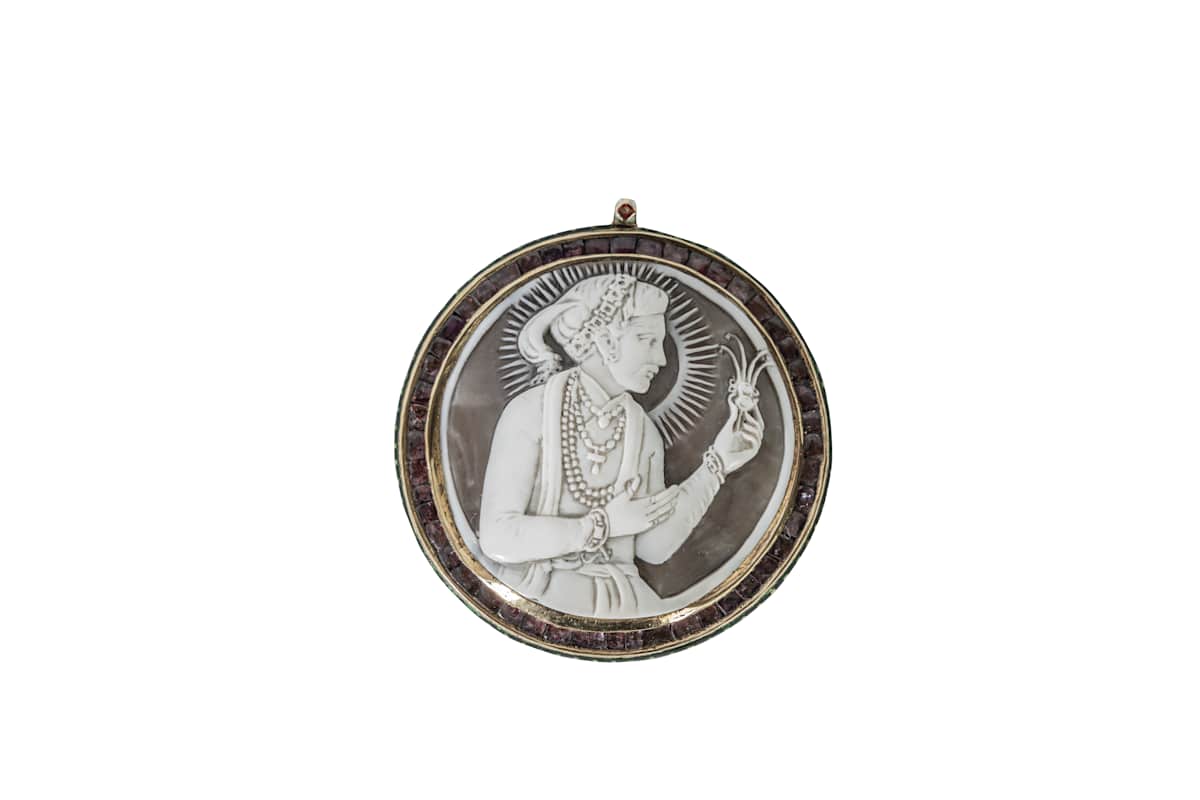 Cameo Pendant of Emperor Shah Jahan  Image: This shell cameo of Shah Jahan, likely carved by a European artist at court, shows the emperor in profile with regal adornments and a divine halo. Framed in rubies, the reverse features vibrant enamelling. Such portrait pendants, introduced by Akbar, were gifted to favored courtiers.