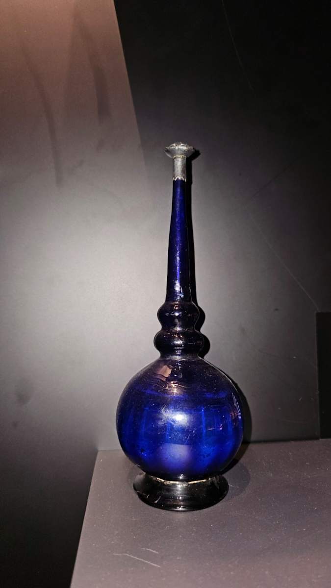 Mughal Rosewater Sprinkler  Image: Blown from deep cobalt-blue glass, this rosewater sprinkler stuns with its globular body, flared foot , and tapering neck ringed in delicate mouldings. A Silver-mounted mouth crowns the vessel, once used to release scented drops in moments of courtly ritual.