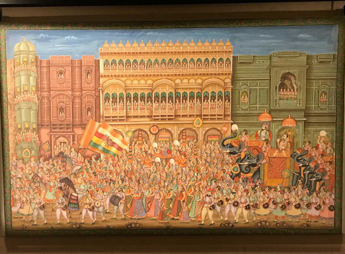 Royal Procession, Marwar  Image: .This monumental painting portrays a grand Rajput procession in Marwar, led by nobles on elephants and cavalry, with vibrant bands of dancers, musicians, and flag-bearers. The flag of Jodhpur, bearing the royal insignia of Marwar, asserts regional identity. Intricately detailed havelis and watching figures in jharokhas frame this vivid display of royal spectacle and urban celebration.