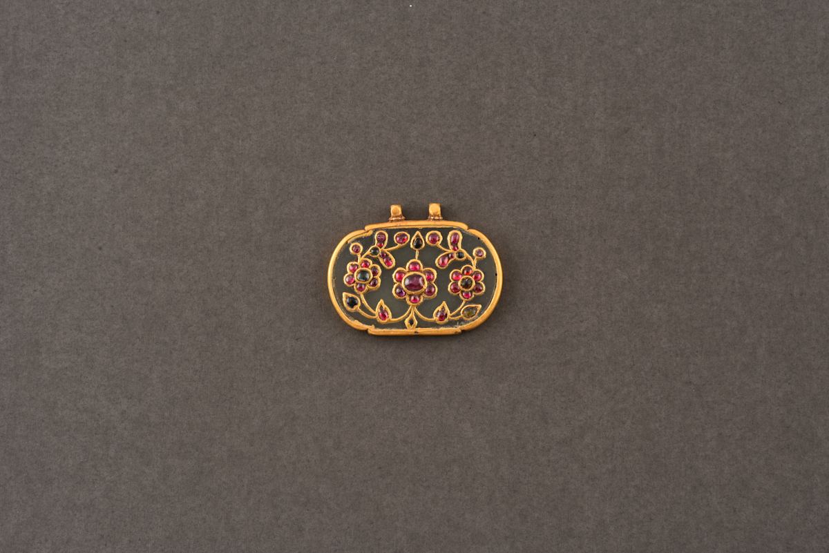 Jade pendant (Haldili)  Image: Carved from translucent 'mutton fat' nephrite jade, this refined Mughal-era pendant features a floral spray inlaid with gold and set with emeralds and rubies in delicate kundan. Beyond its beauty, the pendant was believed to offer talismanic protection and good fortune.
