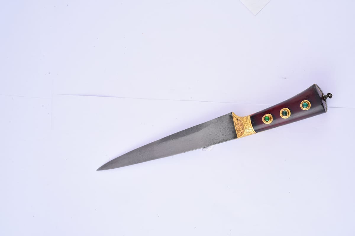 Pesh kabz Dagger  Image: Wootz steel blade with subtle patterns and a strong “T” spine for piercing mail armor, tapering to a fierce triangular point. The hilt features red glass inlays, with gold accents near the base, blending deadly function with regal ornament.