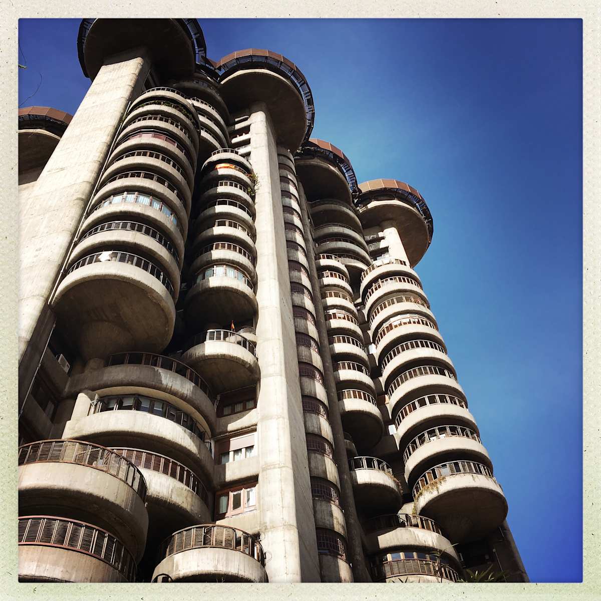 Blue Sky Brutalism by Sherry Ross  Image: A towering Brutalist apartment complex rises against the brilliant blue Madrid sky, its monolithic presence both imposing and mesmerizing. The warm gray concrete, textured and unyielding, absorbs the sunlight, casting deep geometric shadows that emphasize its stark, functional design. Each rigid line and angular form stands in bold contrast to the sky’s vivid expanse, a juxtaposition of heaviness and openness. Despite its raw, unadorned aesthetic, there’s a quiet beauty in its brutal honesty—an architectural statement of permanence and purpose, etched against the endless blue.