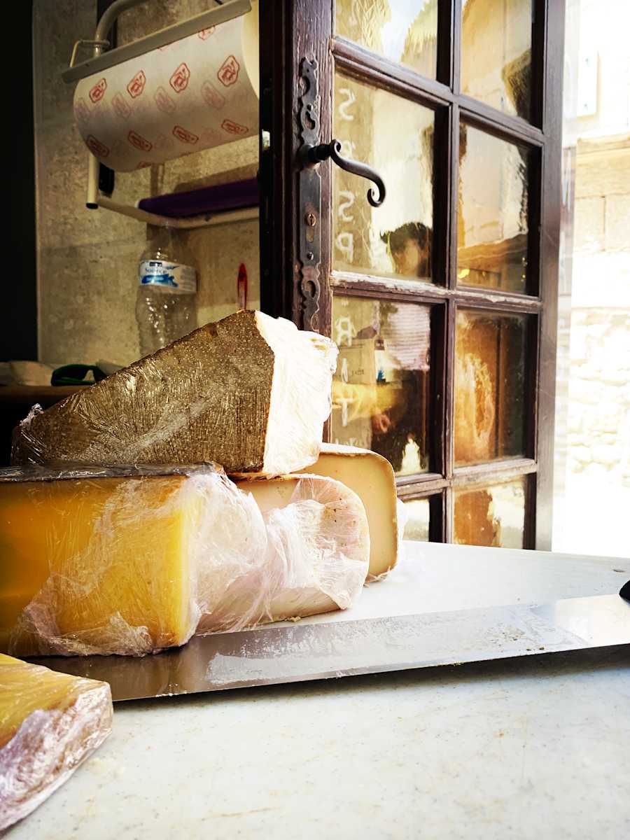 Cheese Shop Window by Sherry Ross  Image: Tucked along a narrow, single-lane cobblestone street in the heart of Carcassonne, a small gourmet shop beckons with the rich aromas of handmade cheeses and briny olives. Within the towering medieval walls, time seems to blur—tourists pause to admire the historic charm while locals move with quiet familiarity, their steps echoing those of shoppers who once strolled these same streets centuries ago. The past lingers here, woven into the stone and the daily rituals of life, where tradition and modern indulgence coexist in the shadow of history.

