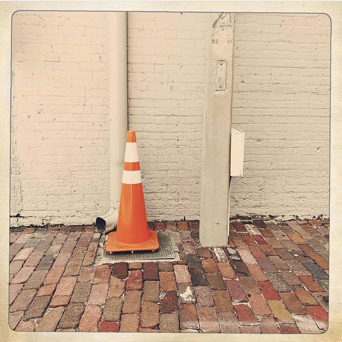 Lone Cone by Sherry Ross  Image: Wandering through the quiet back alleys, I stepped into a scene both unassuming and oddly captivating. A monochromatic brick wall and weathered pole stood as silent sentinels, their layers of paint telling stories of reinvention and time’s persistence. Beneath them, the well-worn brick street stretched out in muted tones, its uneven surface bearing the weight of years, each brick a quiet testament to footsteps past.

Yet, amidst this subdued palette, a single bright orange cone perches squarely atop a grate—its presence both unexpected and absurd. The contrast is stark, almost comical, a small rebellion against the otherwise restrained composition. This simple, accidental arrangement always makes me smile, a quiet reminder that sometimes, humor and beauty emerge in the most unintentional places.