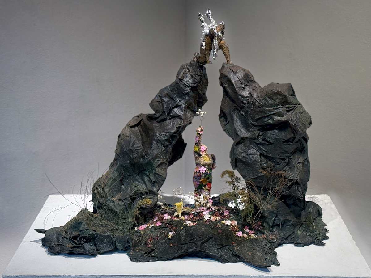 Strength and Vulnerability by Tashina Marie  Image: This sculpture captures the tension between self-protection and openness, strength and vulnerability. At the top, an armored figure, encased in an iron shell of spikes, hovers precariously without support, embodying the weight of guardedness and the illusion of power. Below, a vibrant, flower-covered figure stands firm on solid ground, reaching up with a delicate offering of life and love. Her wings, feathered and light, remain unburdened, symbolizing the true strength found in openness. The piece explores the paradox that shielding oneself from pain also isolates and destabilizes, while embracing vulnerability allows for growth, connection, and true resilience.