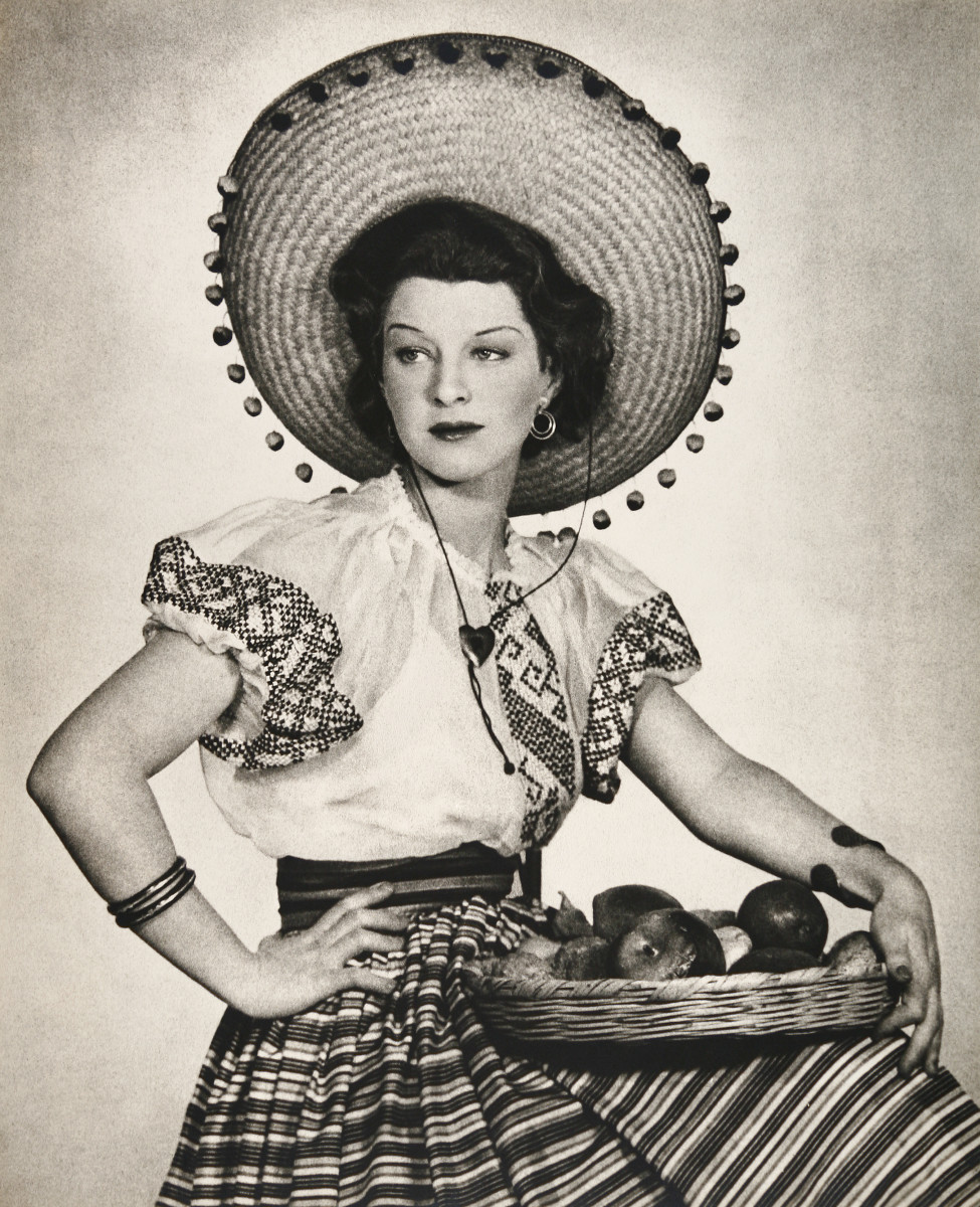 Una Gitana de Mexico by Shirley Hall  Image: A black and white portrait of a seated woman. She is wearing traditional Mexican hat and dress and she has a woven basket of vegetables resting on her lap. 