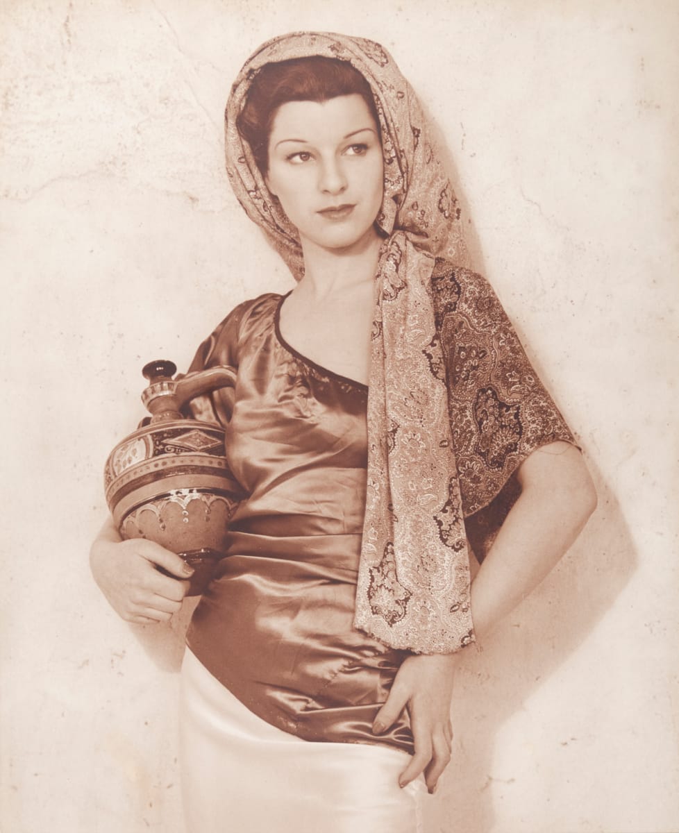 Coquette by Shirley Hall  Image: A sepia toned portrait of a woman standing against a wall. She is wearing a paisley headdress, silk top and holding a decorative jug. 