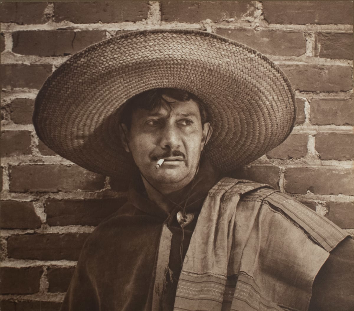 Peon by Shirley Hall  Image: A red-toned portrait of a Hispanic man who is leaning against a brick wall and smoking a cigarette. 