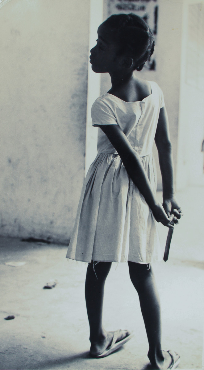 Panama City, Oct. 1963 by Edward R. Miller  Image: A young black child in a white dress faces away from the camera with their hands behind their back and face twisting towards the left of the camera.