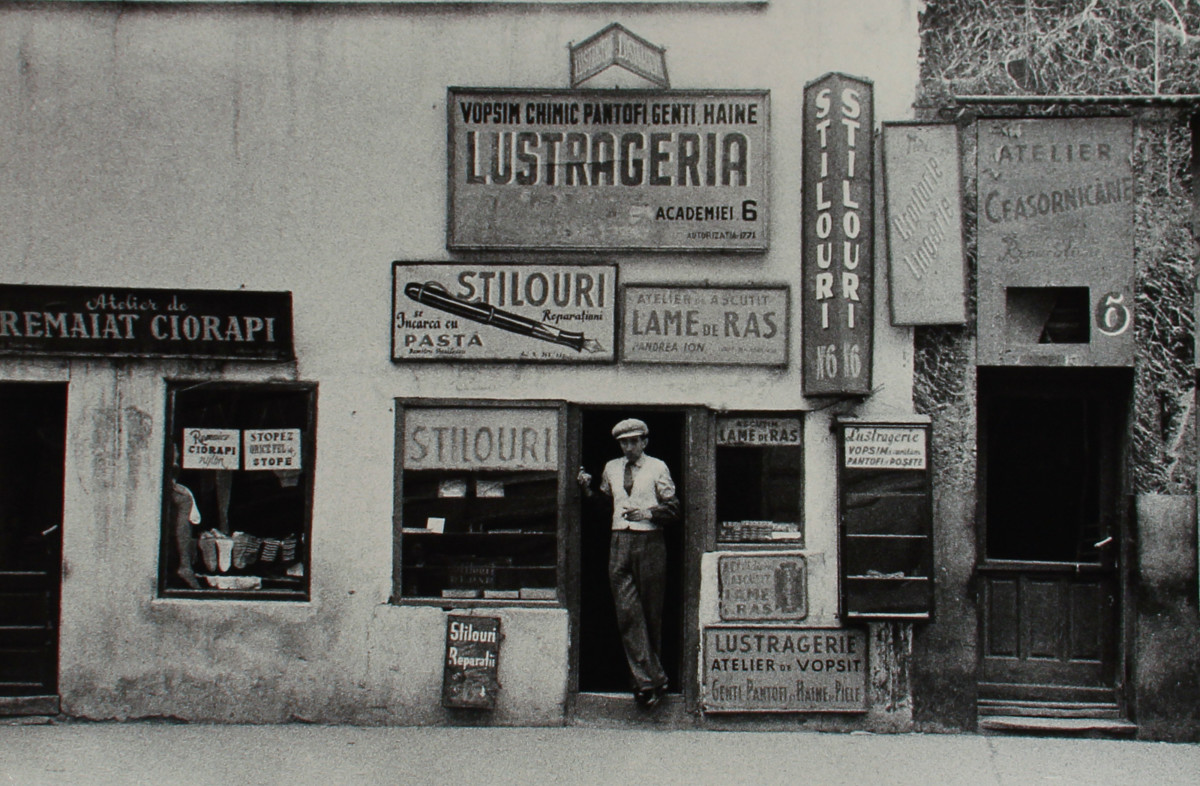 Bucharest, Romania 1956, Storekeeper and Signs by Edward R. Miller  Image: A black and white photograph of a man standing the archway of a storefront.