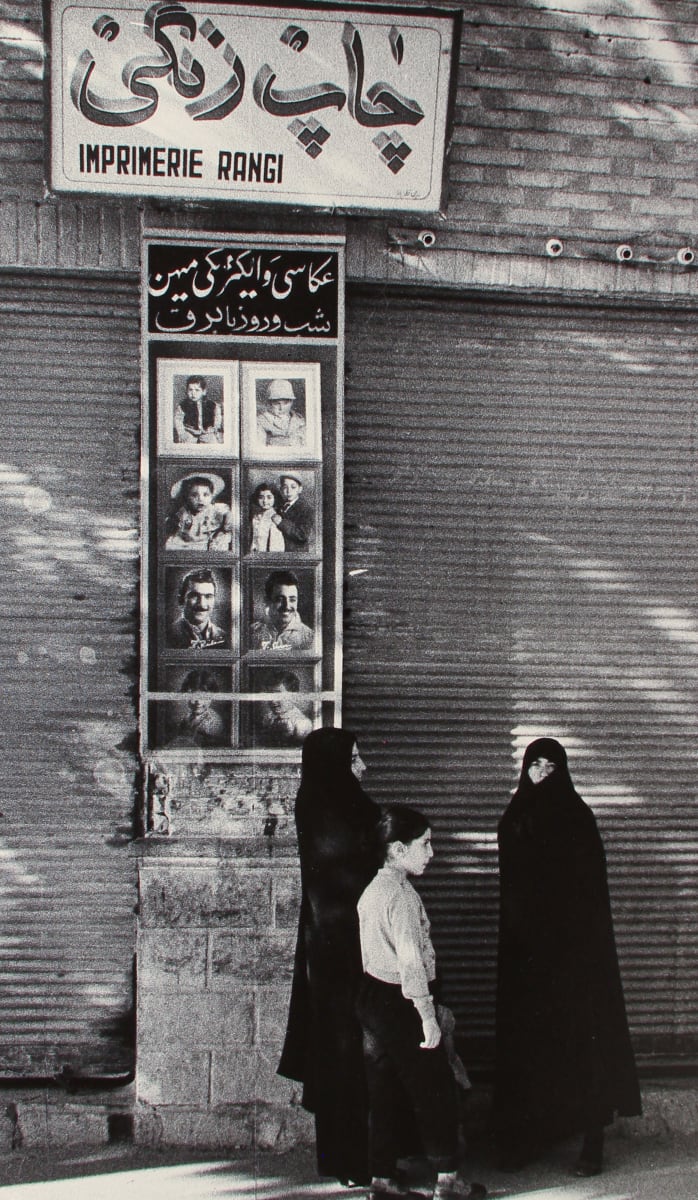 Tehran, Iran 1956 by Edward R. Miller  Image: A black and white photograph of two Muslim women in traditional chadors and a young child in a button up and trousers standing in front of stone pillar.