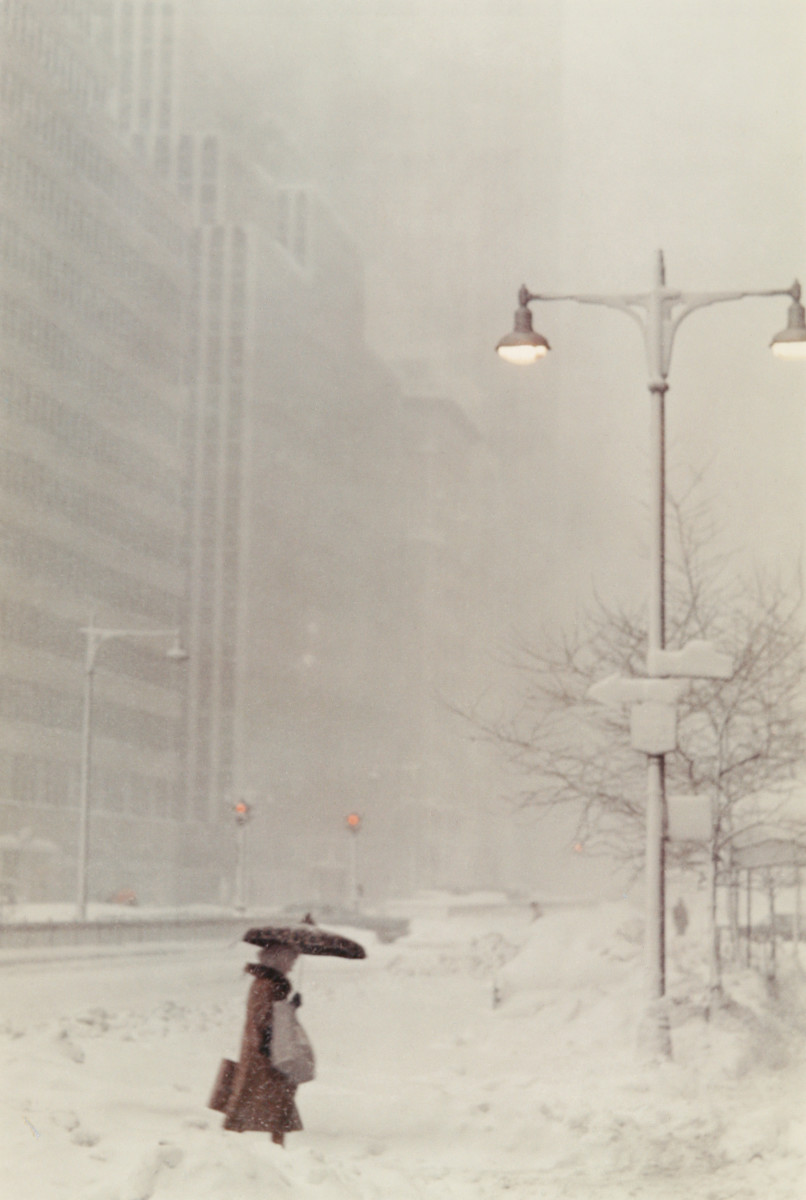 Snowy Day in Manhattan by H. Landshoff  Image: A color photograph of a woman walking through a snowy street. She is carrying two large backs and holding an umbrella over her head. 

