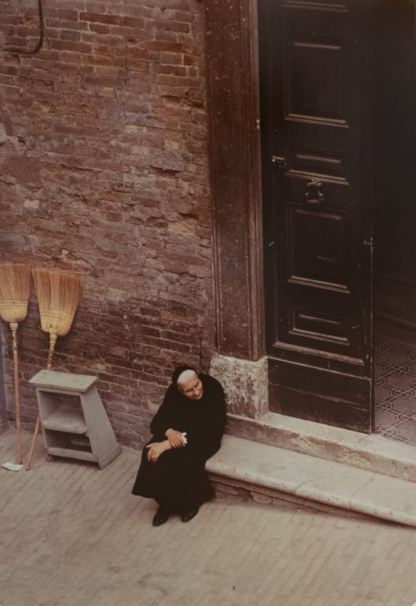 Old Lady on Doorstep by H. Landshoff  Image: A color photograph of an elderly woman in all black sitting on a stoop outside of a brick building.