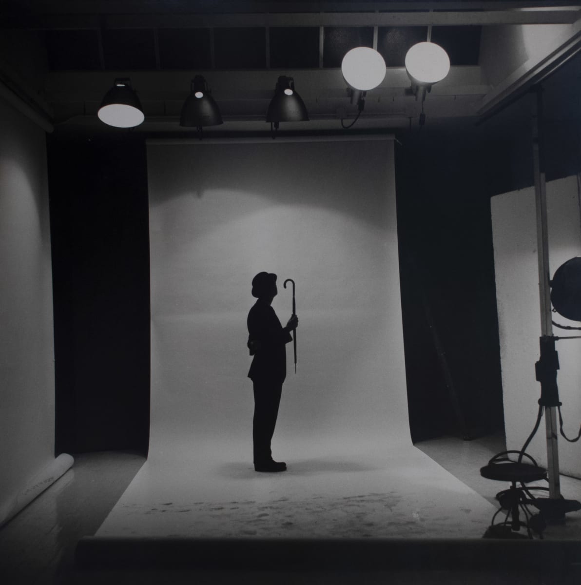 The Umbrella by H. Landshoff  Image: A black and white photograph of a man in standing on a studio backdrop holding a closed umbrella.