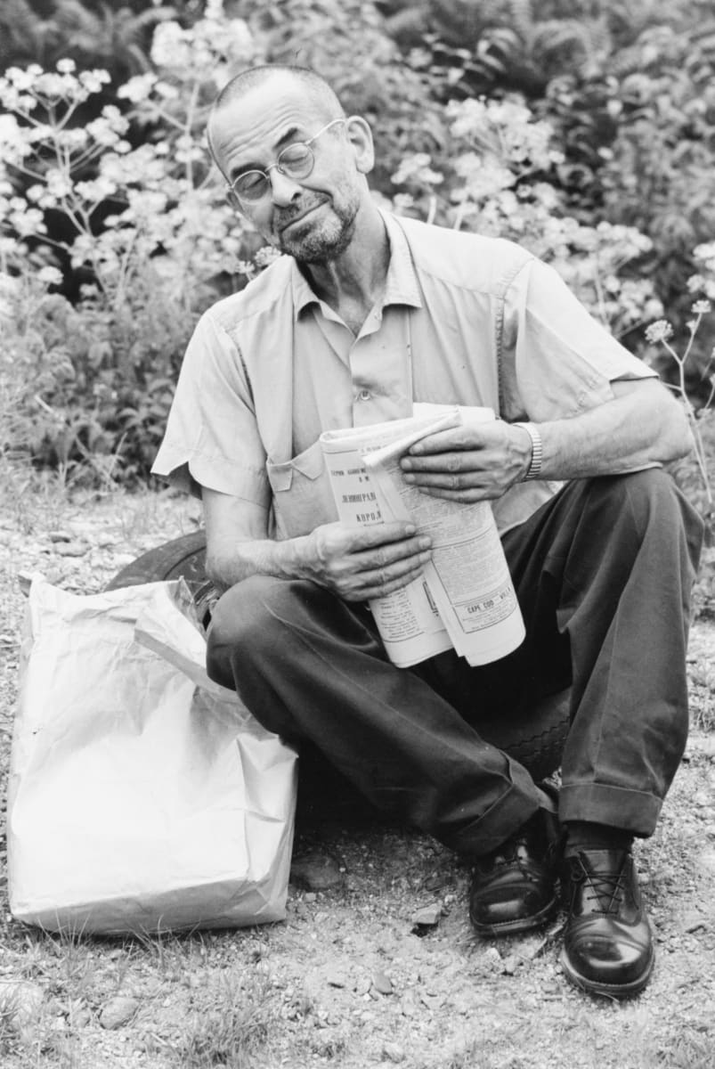 Dr. Cutler West by H. Landshoff  Image: A black and white photograph of a man sitting on gravel. He is wearing glasses and reading a folded newspaper. 