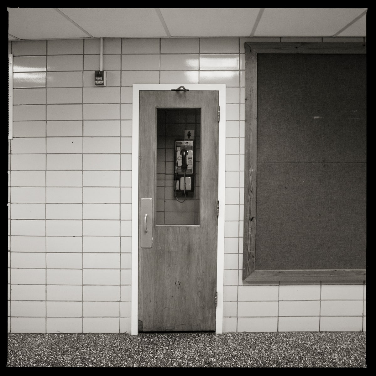 585.223.9793 – Johanna Perrin School, 85 Potter Place, Fairport, NY 14450 by Eric T. Kunsman  Image: ID: A black and white image shows a tiled wall with a door in the middle and a bulletin board to the right. The door has a window on it, and through the window is a payphone.