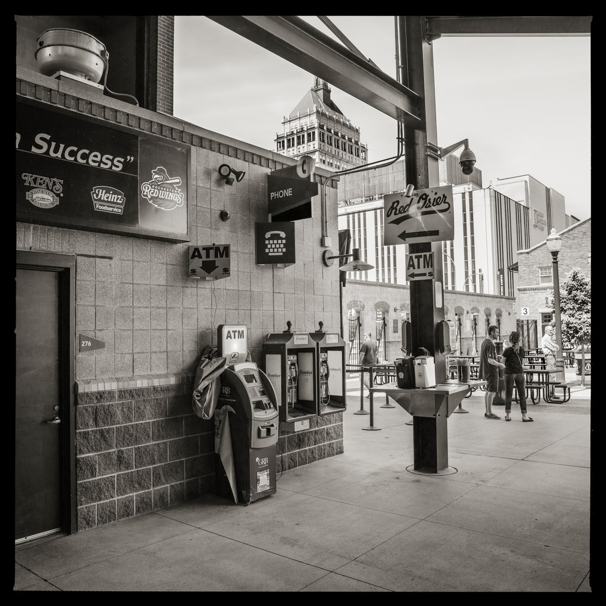 585.232.9300 & 585.232.9437 – Frontier Field, 333 N. Plymouth Ave., Rochester, NY 14608 by Eric T. Kunsman  Image: ID: A black and white image shows a wall on the left with an ATM and two pay phones.  At the end of the wall people are visible and the Kodak Tower is in the background.