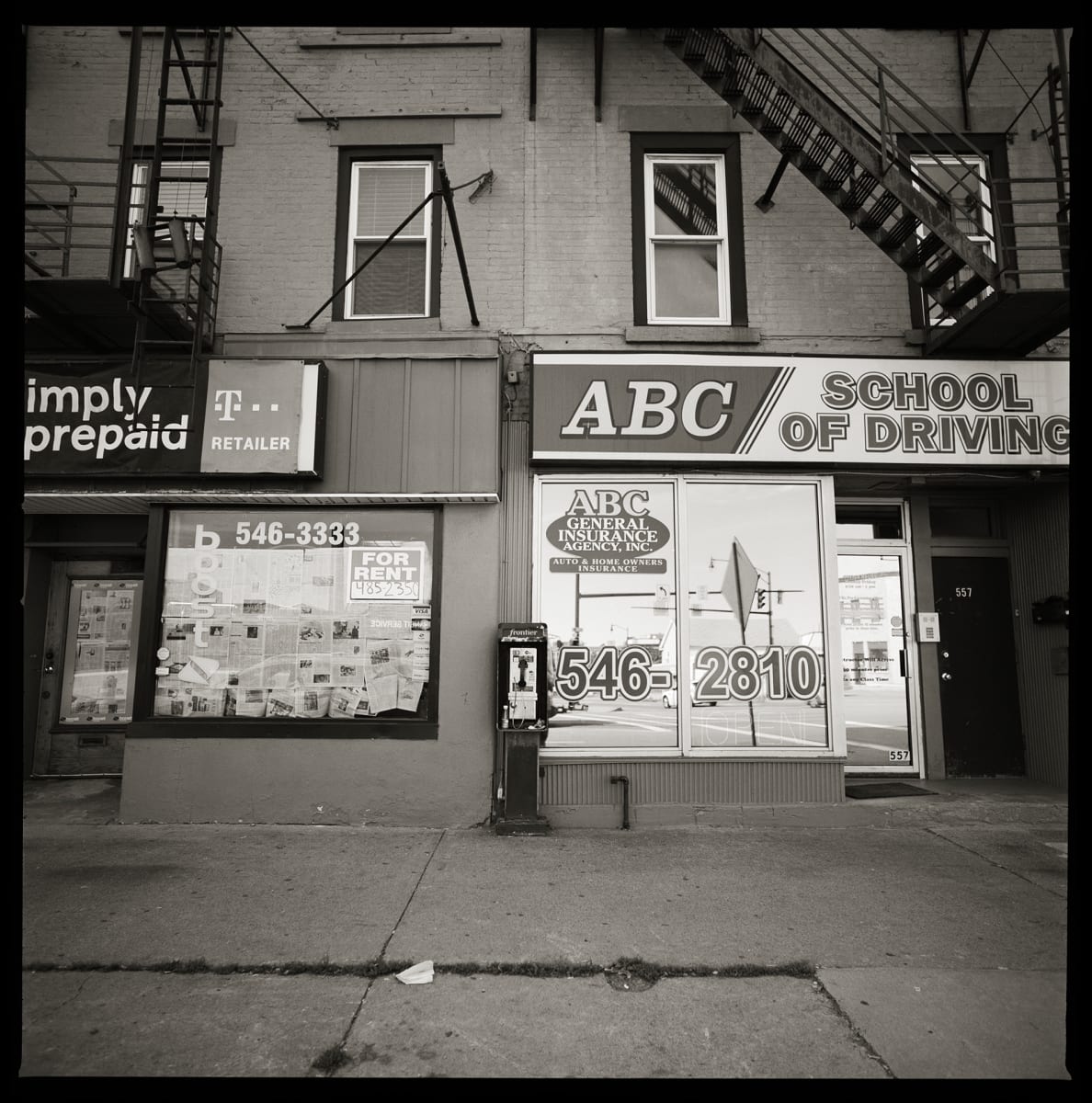 585.232.9384 – ABC School of Driving, 557 State Street, Rochester, NY 14608 by Eric T. Kunsman  Image: ID: A black and white image shows a building with a large glass entrance with advertisements for the ABC School of Driving.  To the left of the entrance there is a payphone.  