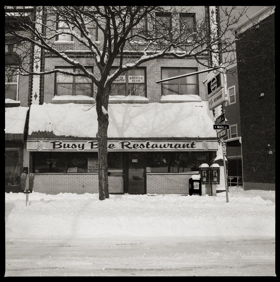 585.232.9542 & 585.232.9262 – Busy Bee Restaurant, 124 West Main Street, Rochester, NY 14614 by Eric T. Kunsman  Image: ID: A black and white image shows a snow covered road and restaurant on a corner.  There are two pay phones on the right side of the restaurant that also have snow on their tops.  The awning says "Busy Bee Restaurant".
