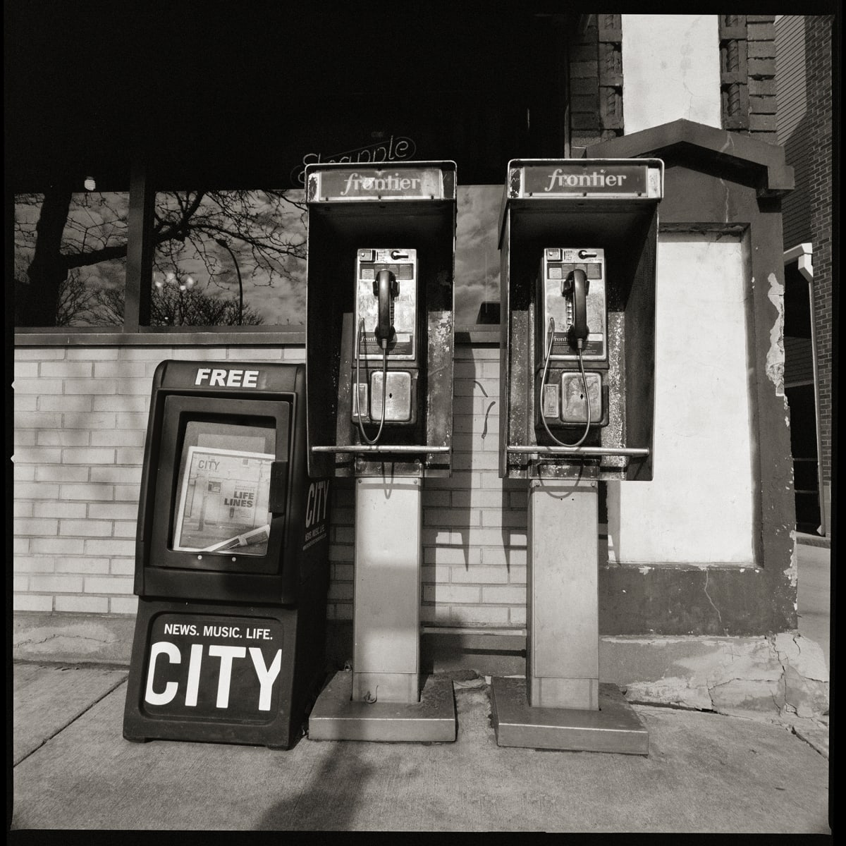 585.232.9542 & 585.232.9262 – Busy Bee Restaurant, 124 West Main Street, Rochester, NY 14614 by Eric T. Kunsman  Image: ID: A black and white image shows two pay phones to the right of a City Newspaper container. This is on a sidewalk.