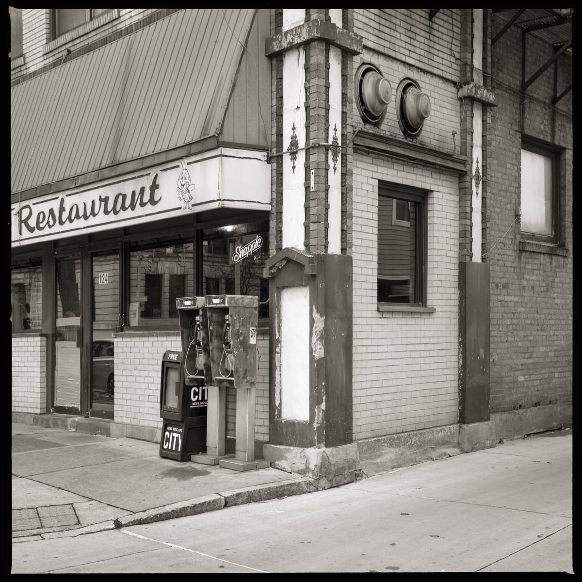 585.232.9542 & 585.232.9262 – Busy Bee Restaurant, 124 West Main Street, Rochester, NY 14614 by Eric T. Kunsman  Image: ID: A black and white image shows a corner restaurant with an awning.  Under there right side of the awning are two pay phones.  The building is surrounded by a side walk.