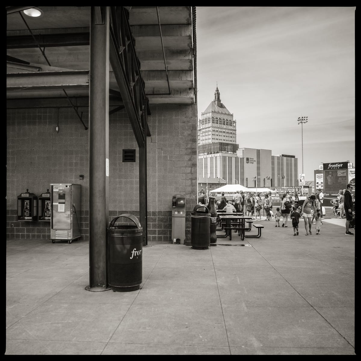 585.232.9882 & 585.232.9300 – Frontier Field, 333 N. Plymouth Avenue, Rochester, NY 14608 by Eric T. Kunsman  Image: ID: A black and white image shows a building overhang on the left and on the right is the Kodak tower and other buildings surrounding it and people in the foreground. Under the building overhang there are two payphones and a garbage can.