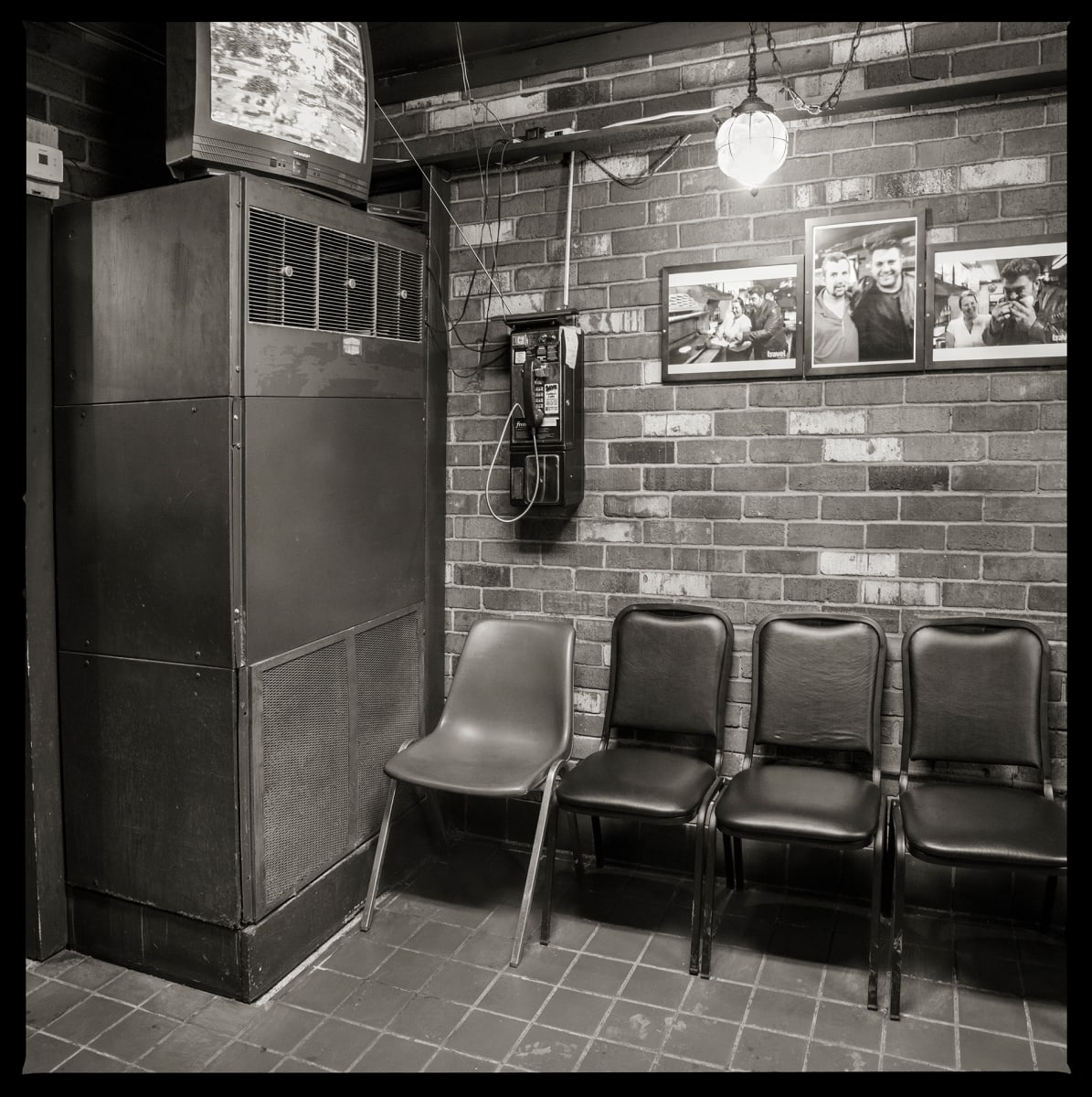 585.235.9156 – Campi’s Restaurant, 205 Scottsville Road, Rochester, NY 14611 by Eric T. Kunsman  Image: ID: A black and white image shows a metal cabinet against a left side of a corner.  On the right side of the corner is a brick wall with four chairs against it, and a wall-mounted payphone.  To the right of the payphone are a variety of framed images.