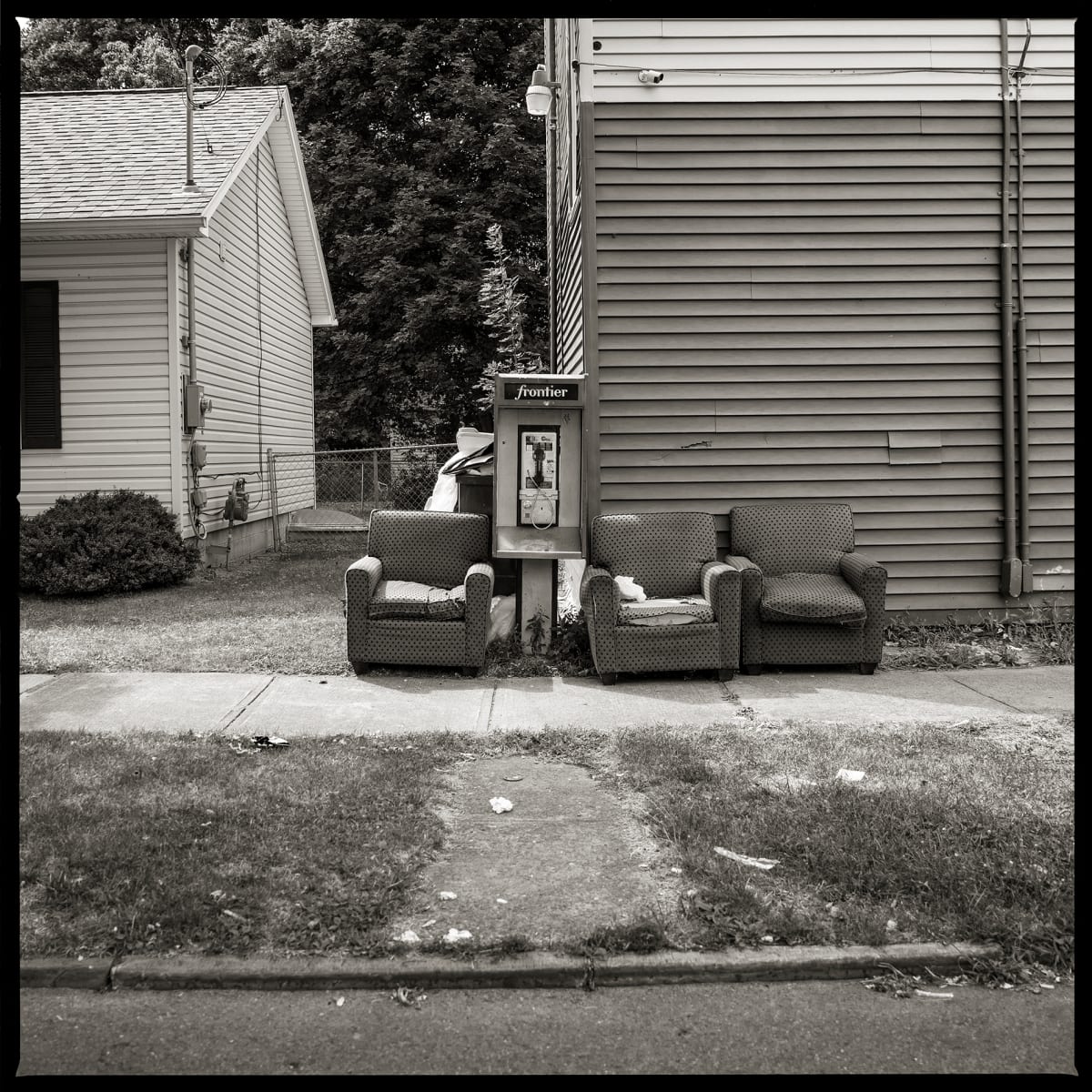 585.235.9196 – Grape and Orange Mini Mart, 111 Orange Street, Rochester, NY 14611 by Eric T. Kunsman  Image: ID: A black and white image shows a payphone in front of two buildings.  Around the payphones are three armchairs behind the side walk.
