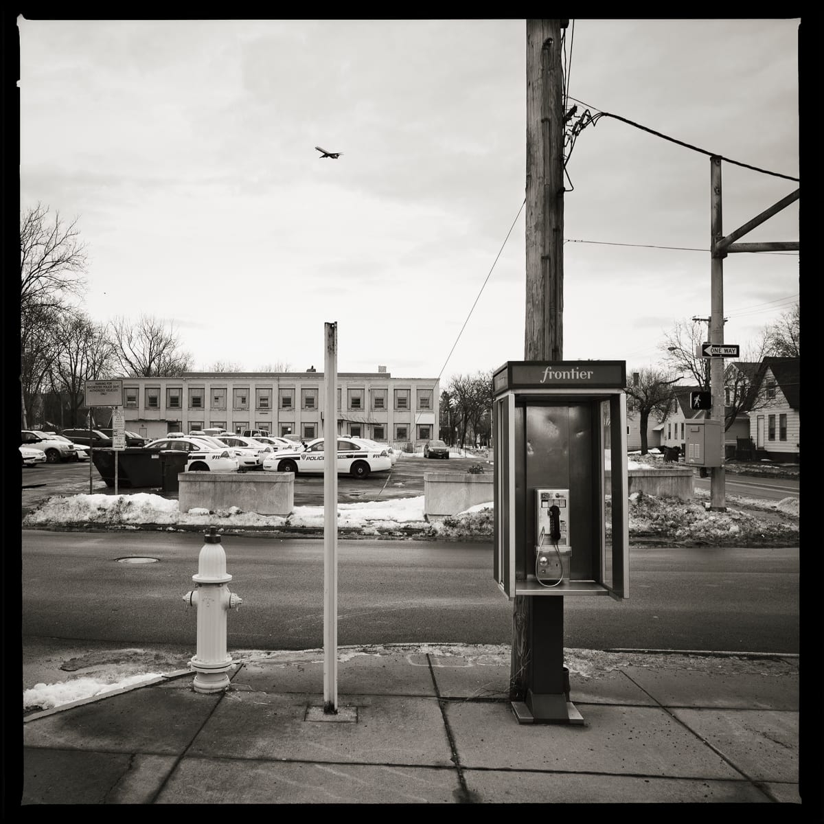585.235.9493 – Rochester Police Department, 261 Child Street, Rochester, NY 14611 by Eric T. Kunsman  Image: ID: A black and white image shows the Rochester police station from across the street.  There are many cop cars in front of the building.  On this side of the road is a payphone next to a telephone pole.  There is a bird flying in the frame as well.