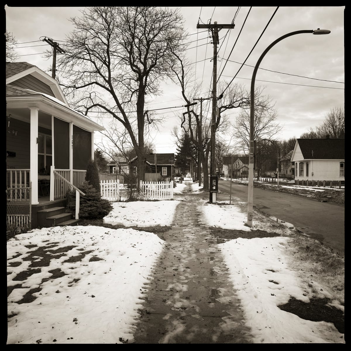 585.235.9583 – 167 Whitney Street, Rochester, NY, 14611 by Eric T. Kunsman  Image: ID: A black and white image shows a snowy sidewalk in a residential area.  Houses are on the left and the road is to the right.  There is a payphone next to a telephone pole to the right of the sidewalk. 