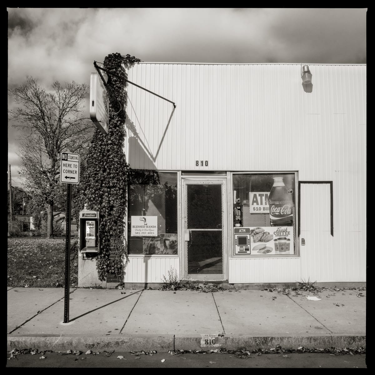 585.235.9596 – Shaibe Grocery, 810 Brown Street, Rochester, NY 14611 by Eric T. Kunsman  Image: ID: A black and white image shows a white building that has a center entrance.  The left of the entrance is covered in ivy, and in the ivy is a payphone.