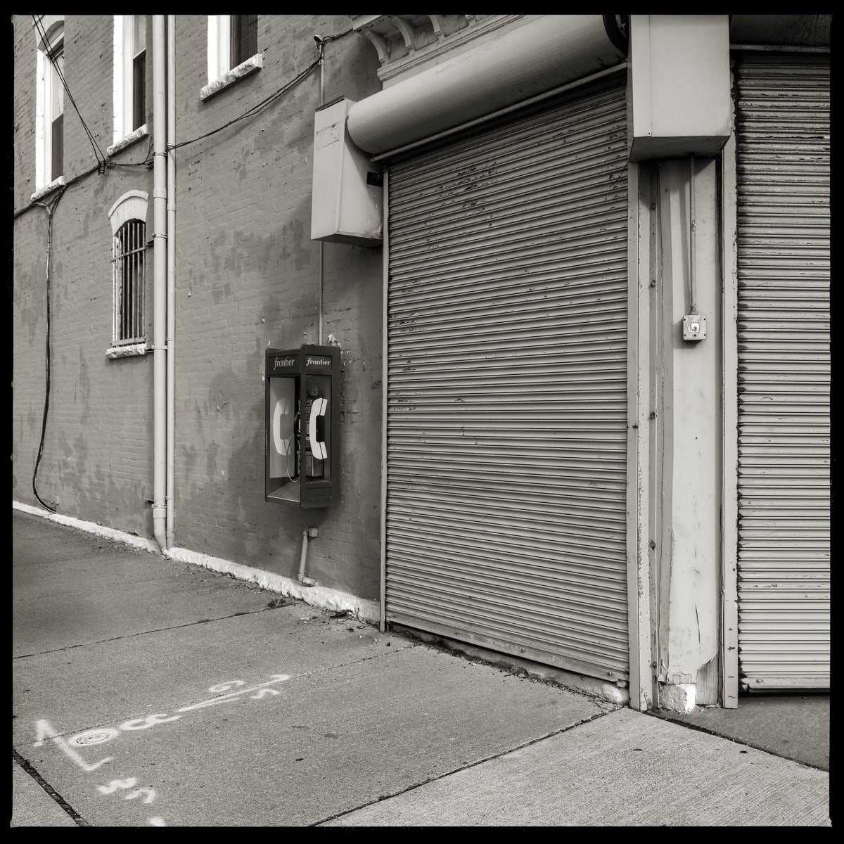 585.254.9463 – Aladdin’s Communications, 1108 Lyell Avenue, Rochester, NY 14606 by Eric T. Kunsman  Image: ID: A black and white image shows a building that is partially shut up with a garage-like door. To the left of this door is a payphone mounted to the brick wall.  There are windows to the left of the payphone.