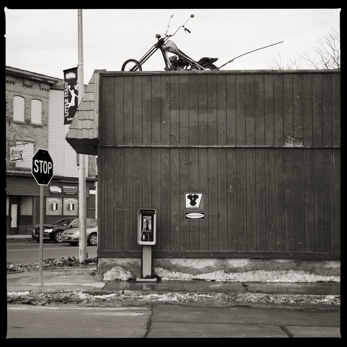 585.254.9533 – Freebird Cycles, Lyell Avenue, Rochester, NY 14618 by Eric T. Kunsman  Image: ID: A black and white image shows a payphone against a dark grey building on a corner.  On the roof of the building is a motorcycle statue.  There is a stop sign to the left.