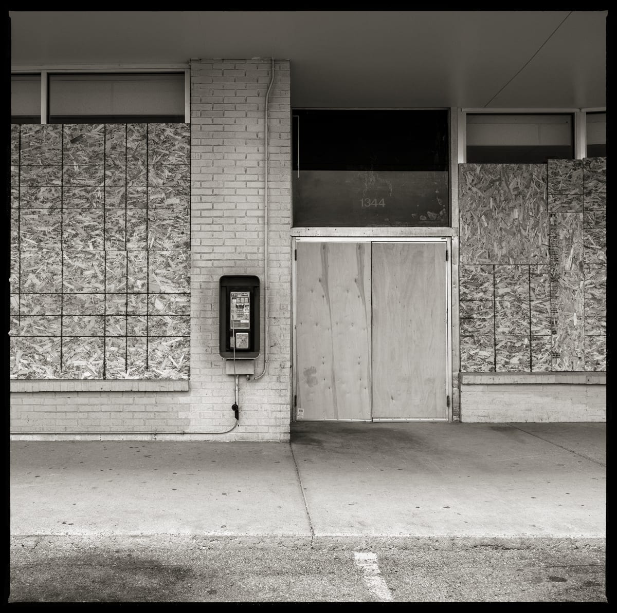 585.254.9782 – Family Dollar Store #1962, 1340 Lyell Avenue, Rochester, NY 14606 by Eric T. Kunsman  Image: ID: A black and white image shows a boarded up building with a payphone mounted a the brick pillar.