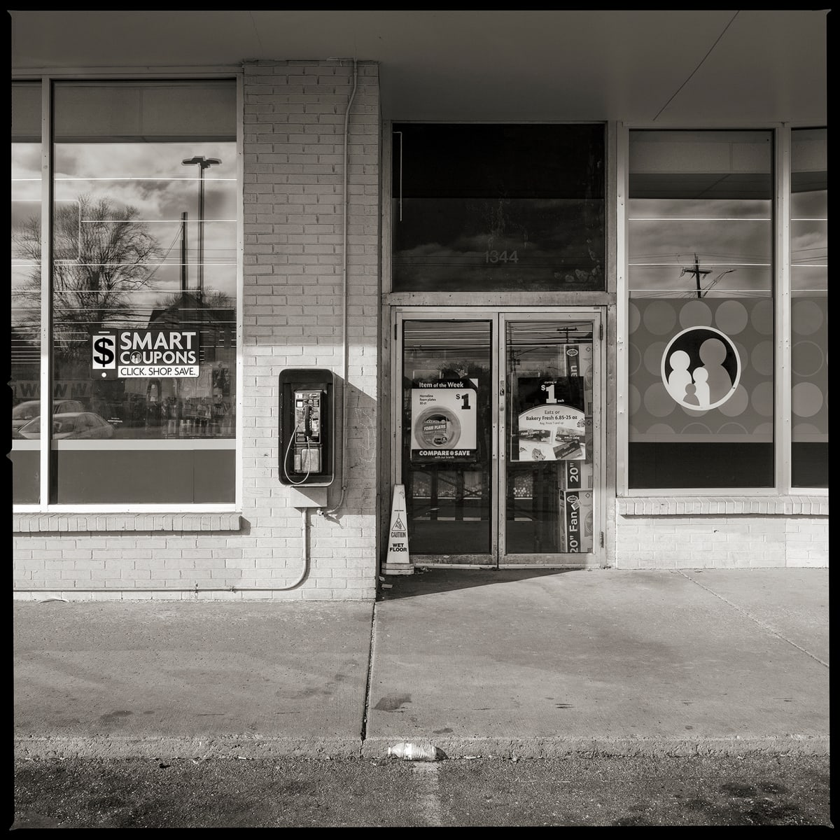585.254.9782 – Family Dollar Store #1962, 1340 Lyell Avenue, Rochester, NY 14606 by Eric T. Kunsman  Image: ID: A black and white image shows a payphone mounted to a storefront.  The left side of the payphone is a window with a Smart Savers advertisement.  The right side of the payphone is the glass door entry way to the building that is covered in advertisements and has a "Caution Wet Floor" sign outside of the door.
