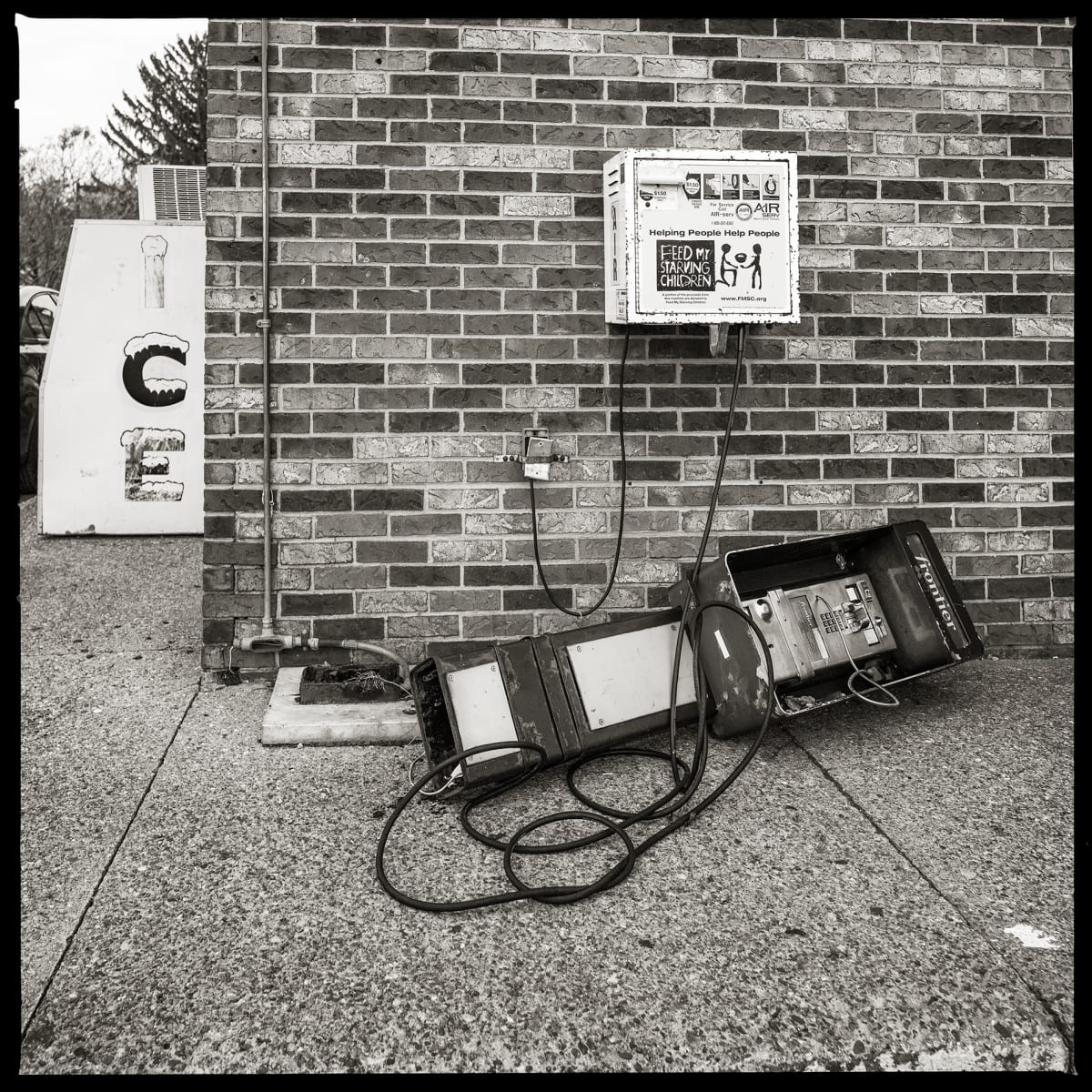 585.266.9507 – AB Convenient Market, 1684 Norton Street, Rochester, NY 14609 by Eric T. Kunsman  Image: ID: A black and white image shows a payphone that has been destroyed.  It is laying on the sidewalk with wires around it.  There is a brick building in the background.