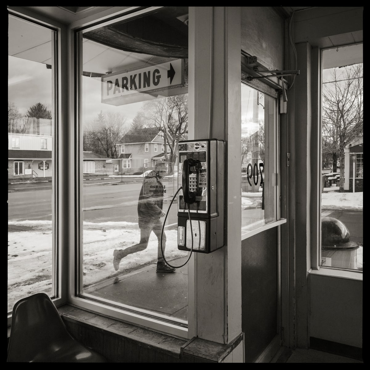 585.266.9536 – RJC Sudsville Laundry, 709 Titus Avenue, Rochester, NY 14617 by Eric T. Kunsman  Image: ID: A black and white image shows a payphone in the middle of a reflective glass entry way. There is a person visible in the window to the left of the payphone.  To the right is the entrance to the building.