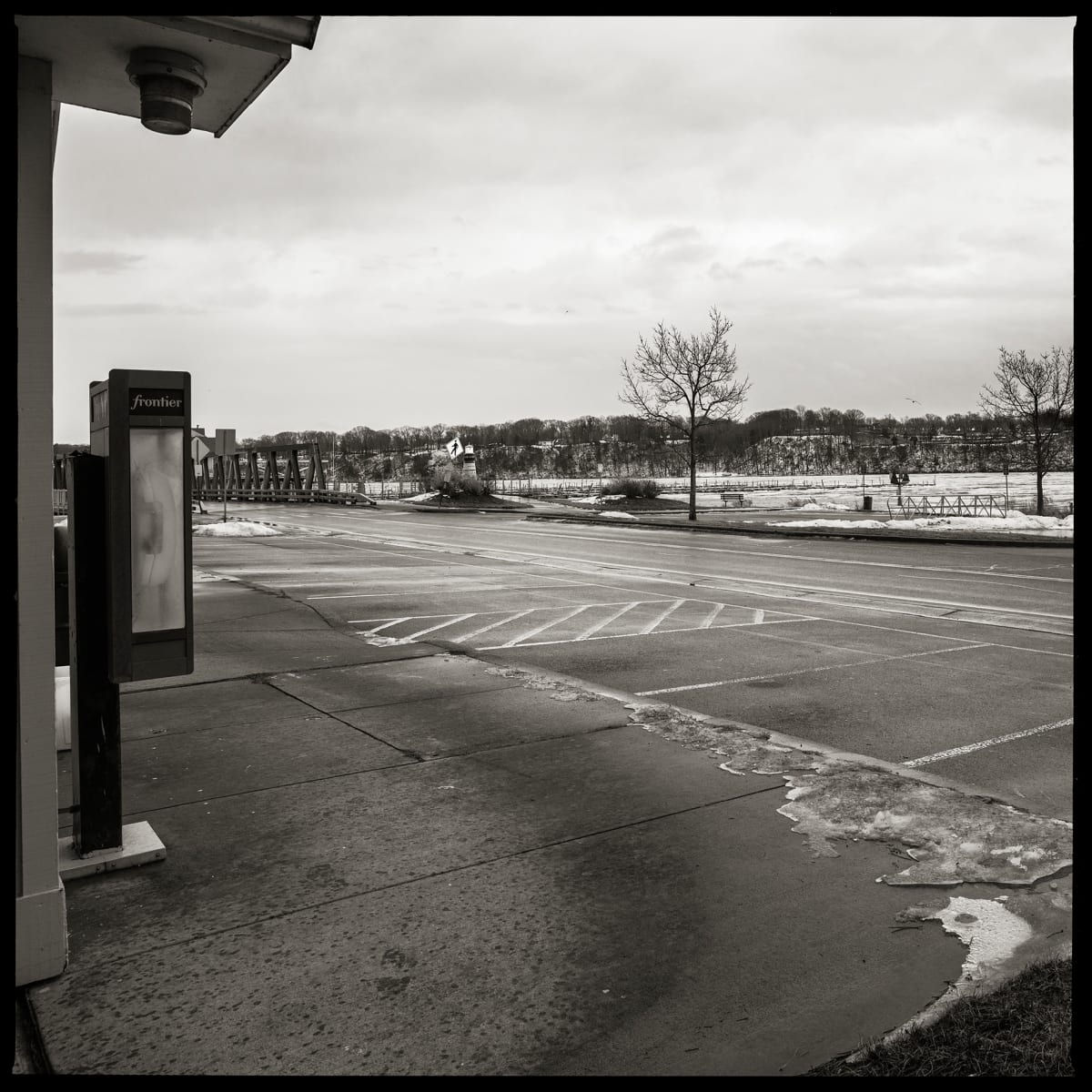 585.266.9557 – Monroe County Parks, 4993 Culver Road, Rochester, NY14622 by Eric T. Kunsman  Image: ID: A black and white image shows an empty parking area with a sidewalk to the left and a snowy field behind.  On the sidewalk is a payphone booth under a building overhang.
