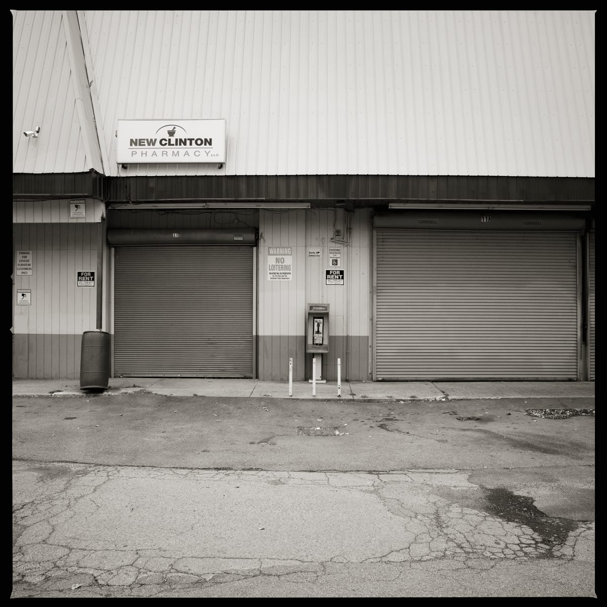 585.266.9867 – Red Wing Plaza, 1415 North Clinton Avenue, Rochester, NY 14621 by Eric T. Kunsman  Image: ID: A black and white image shows two closed grey garage-style doors on a building that holds a pharmacy.  In between the doors is a lighter grey paint on the building and a payphone.