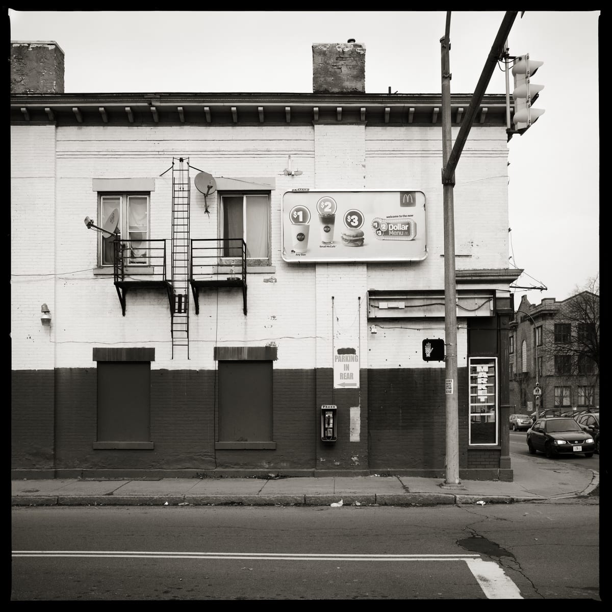 585.266.9901 – Collazo Food Mart, 609 Hudson Avenue, Rochester, NY 14621 by Eric T. Kunsman  Image: ID: A black and white image shows a two-toned building, white on top and dark grey on bottom.  There are various advertisements on the building and a fire exist on the second floor.  There is a payphone mounted on the bottom half of the building.