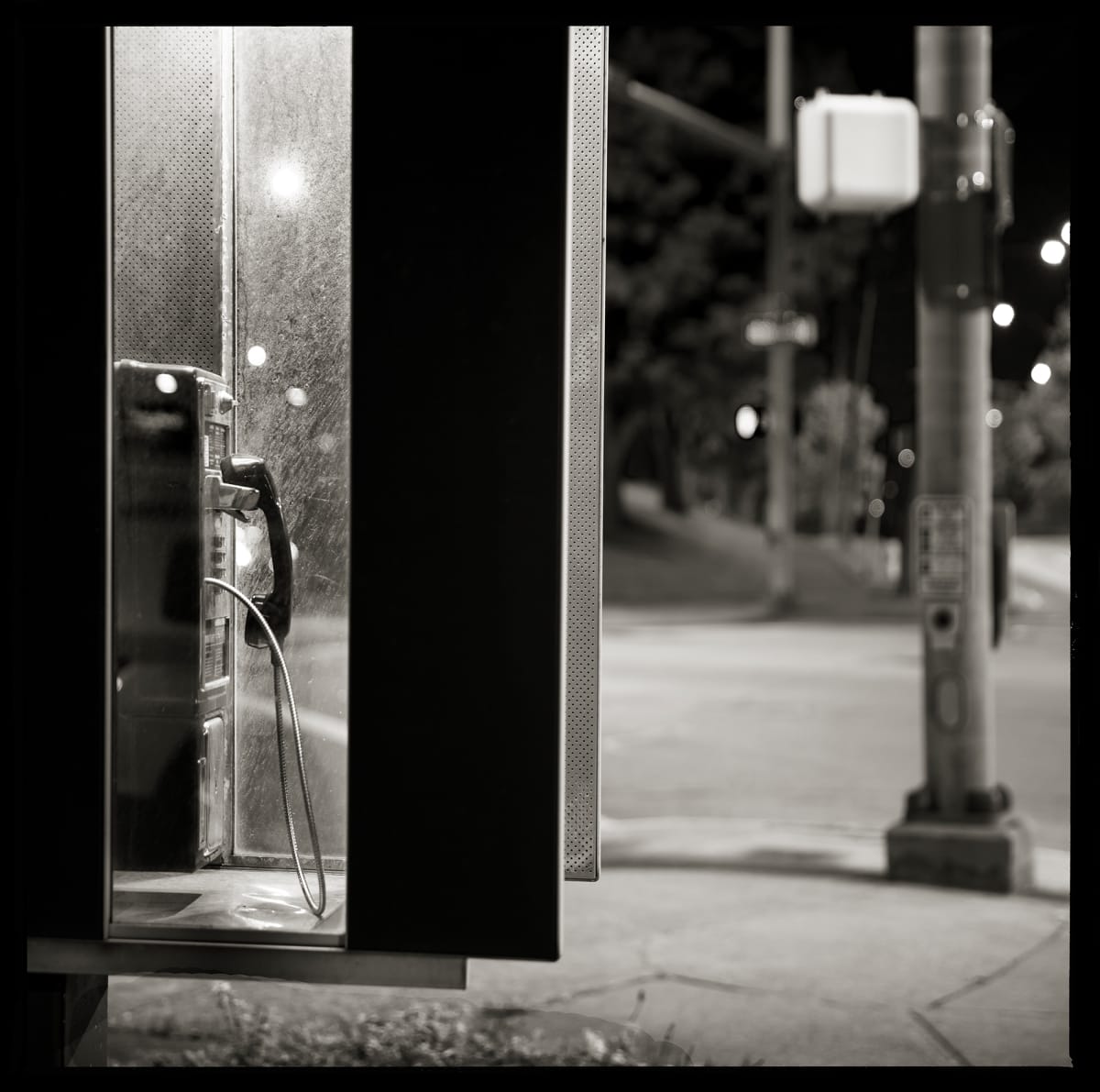 585.271.9197 – Clar’s Elmwood Automotive, 870 Elmwood Avenue, Rochester, NY 14620 by Eric T. Kunsman  Image: ID: A black and white image shows a payphone through the side of the glass booth.  The image is at night, but there is a street light behind the payphone lighting it up.  The payphone is on the left of the image and a telephone pole is to the right.  This is on a street corner.