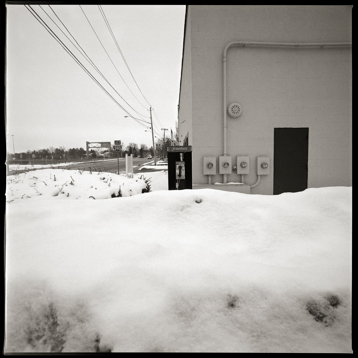 585.271.9607 – Red Apple Market #2025, 1835 Mt. Hope Avenue, Rochester, NY 14620 by Eric T. Kunsman  Image: ID: A black and white image shows a snow covered payphone to the left of a building.  The snow pile is from plowing the parking lot.  The building is a plain grey stucco building.