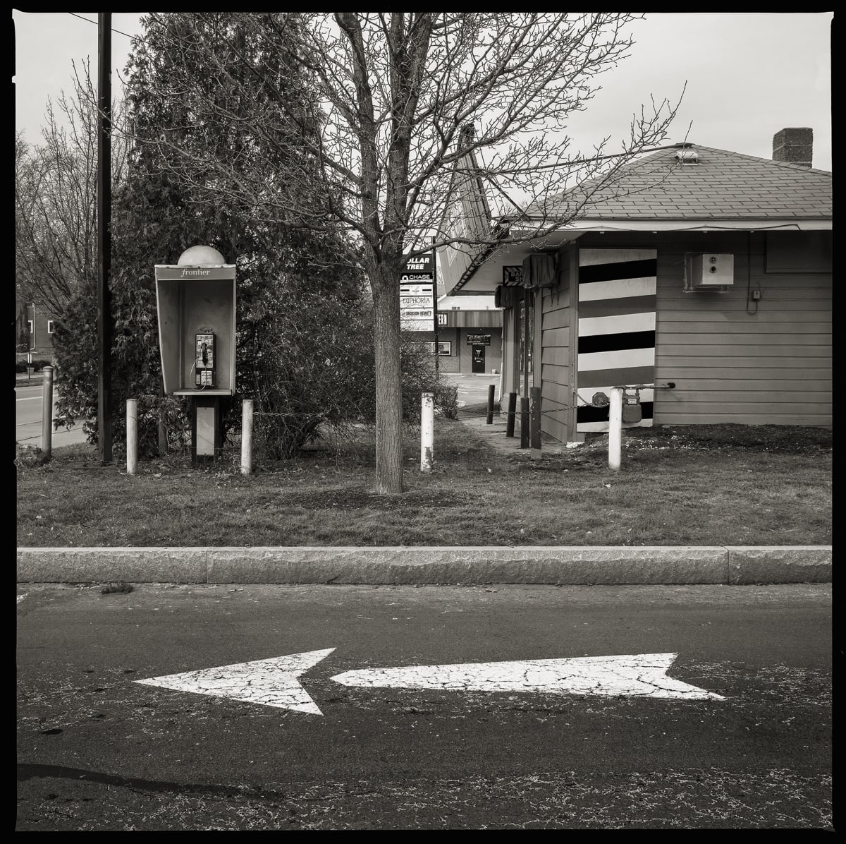 585.288.9518 – 2320 East Main Street, Rochester, NY 14609 by Eric T. Kunsman  Image: ID: A black and white image shows a white arrow in the road pointing left.  Behind the head of the arrow in the grass is a payphone and to the right is a building.