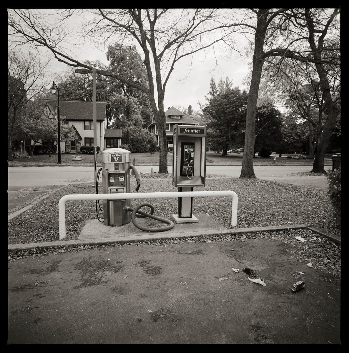 585.288.9865 – Adams Mobile Service, 575 North Winton Road, Rochester, NY 14610 by Eric T. Kunsman  Image: ID: A black and white image shows a payphone next to an air compressor for filling tires.  There is a street behind the payphone.
